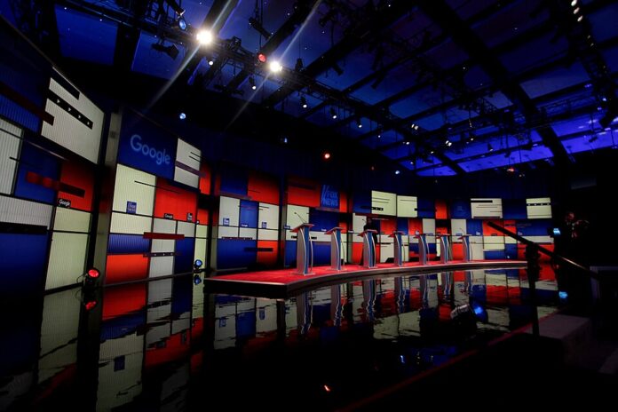 A stage with various red, blue and white squares and seven speaker stands. The stands are empty and the stage's lighting and some camera fixtures are shown.