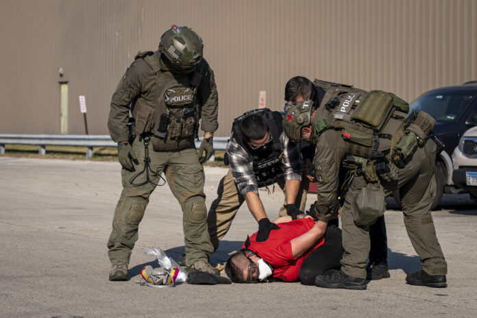 Chicago ICE Operations Four Immigration Control Enforcement agents are shown putting a man in a red shirt in hand cuffs on the floor.