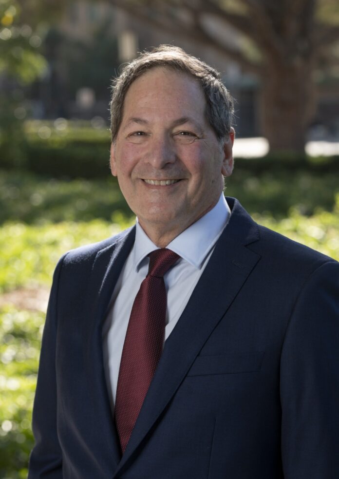 IMG_6722 A portrait of Michael Leon in a red tie and suit are shown. He stands in front of a blurred background of green shrubbery and trees.
