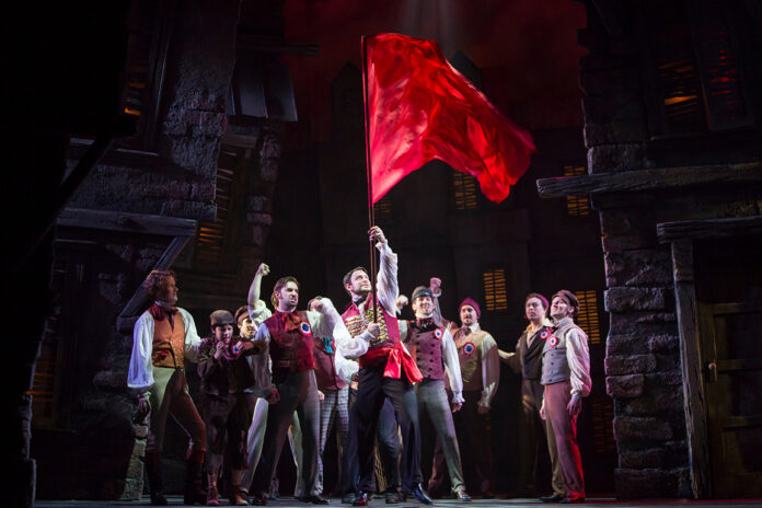 Les_Misérables Castmembers of Les Miserables are shown looking up at a flowing red flag being held up by a man at center stage.