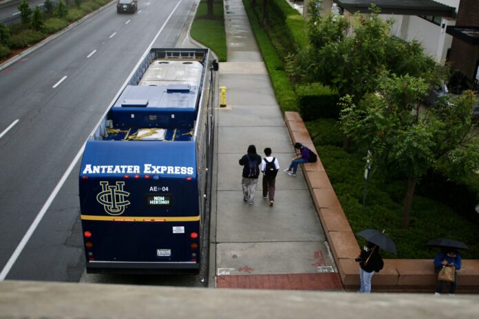 News.UCIpolicesearch.Alex_Mendez_Staff A bird’s eye view of the Anteater Express bus at the University Town Center at UCI is shown parked at the bus stop as students are walking along the side of it, a student is seen with an umbrella.