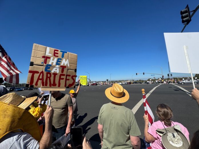 A few protesters are shown at a large intersection with posters and American flags. A poster on the left side reads, "LET THEM EAT TARIFFS."