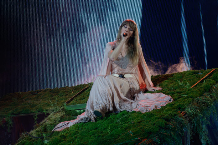 Pop singer Taylor Swift sings on a slanted moss platform wearing a light pink and lace dress. Photo was taken at the Arlington, Texas Eras Tour concert.